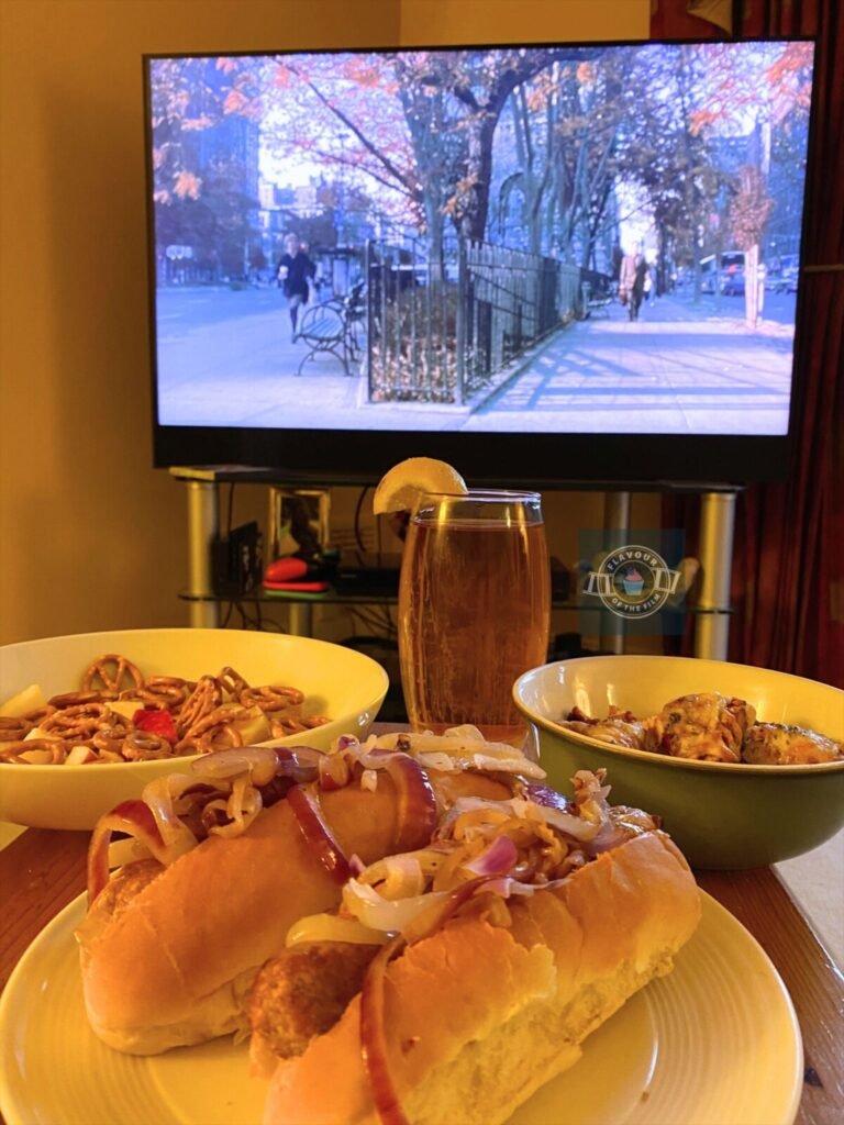 Pretzel snacks with apple chunks, iced tea, a pizza bagel cut up into pieces and two hot dogs topped with fried onions and ketchup plated up and displayed in front of a TV playing You've Got Mail.