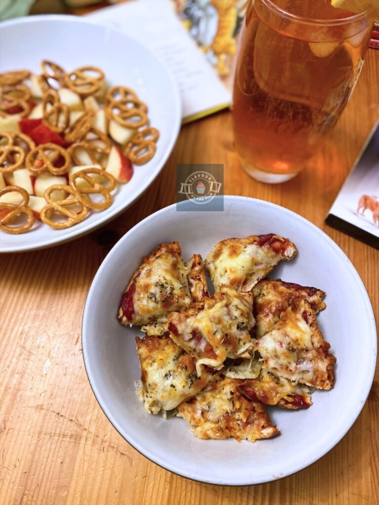 Pizza bagel pieces displayed in a bowl next to a glass of iced tea and dish of pretzel snacks and chopped apples. Props include a DVD copy of You've Got Mail, a white rose and a pile of Ladybird children's books.