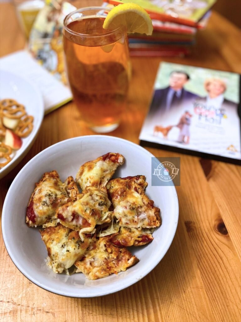 Pizza bagel pieces displayed in a bowl next to a glass of iced tea and dish of pretzel snacks and chopped apples. Props include a DVD copy of You've Got Mail, a white rose and a pile of Ladybird children's books.