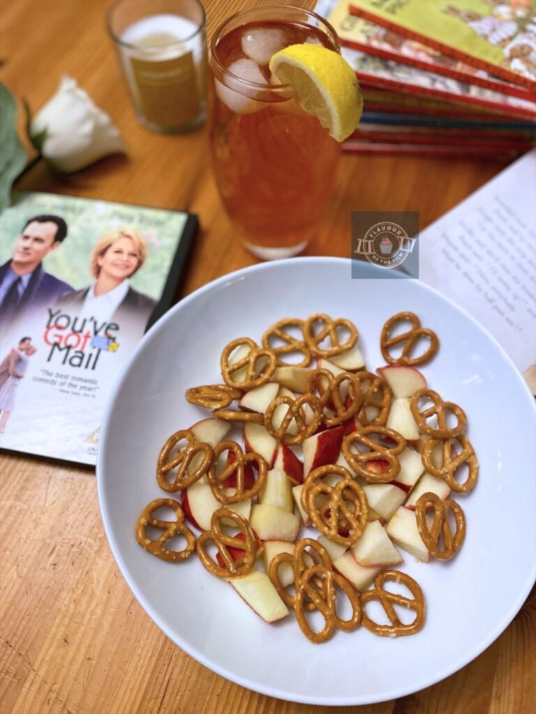 A dish of pretzel snacks and chopped apples next to a glass of iced tea. Props include a white rose, a candle, a DVD copy of You've Got Mail and a pile of children's Ladybird books.