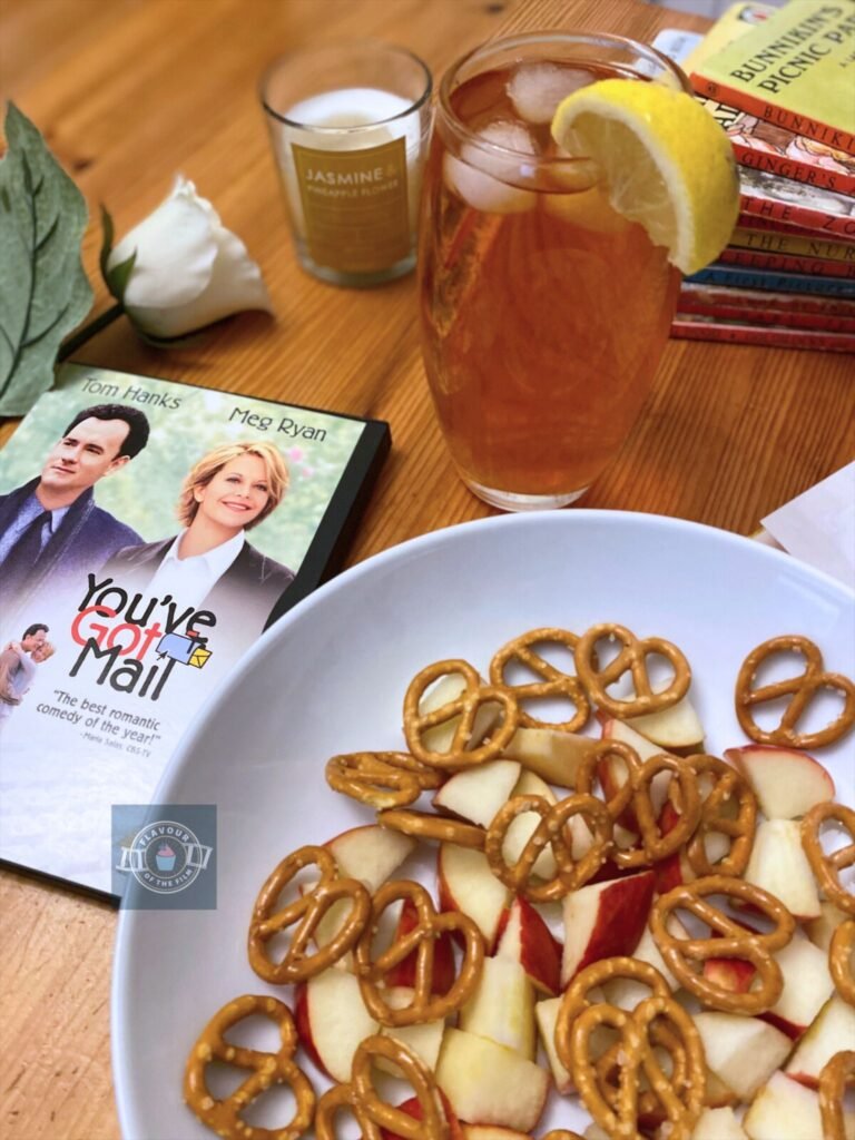 A dish of pretzel snacks and chopped apples next to a glass of iced tea. Props include a white rose, a candle, a DVD copy of You've Got Mail and a pile of children's Ladybird books.