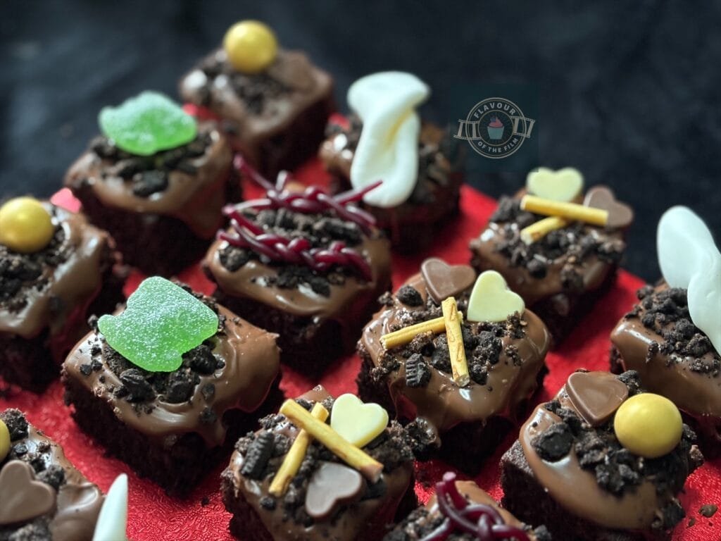 All images are of bite sized chocolate cake squares topped with set melted chocolate, Oreo crumb pieces and varying Halloween themed candy and chocolate elements like hearts. The cake bites are displayed on a red square cake board. Props include mini poison bottle earrings, a Halloween dog jumper with "Boo!" written on it and a pair of white heels.