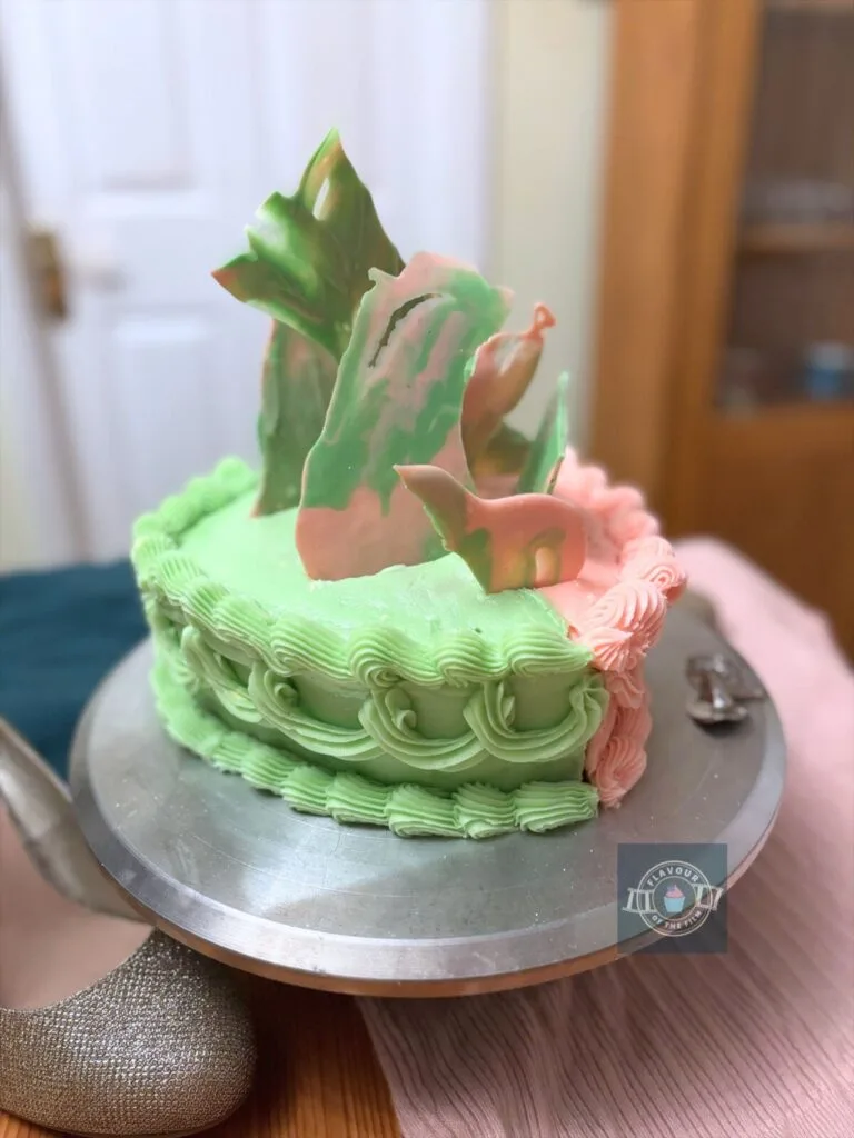 All images are of a two layer heart shaped cake - one half is green and one half is pink. The cake is decorated with green buttercream and pink buttercream, one colour for each half. The cake has a piped top and bottom border and bunting-style piping around the side. There are pink and green chocolate shards protruding from the top of the cake like frozen magical spells. The cake is presented on a silver stand and props include a silver witch hat Wicked keyring, a pair of glittery gold shoes, green material and pink material.