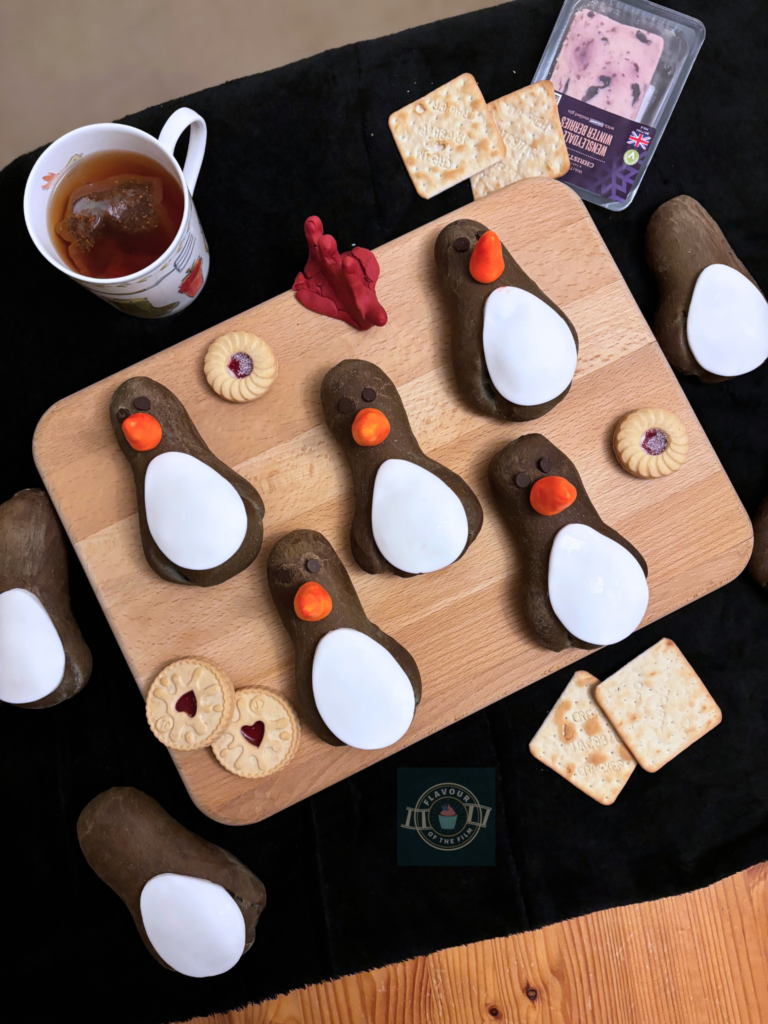 All images are of bread buns in the image of Feathers McGraw aka the villainous penguin from two Wallace and Gromit films. The buns are displayed on a wooden cheese board. Props include a cup of tea, some biscuits, Wensleydale cheese and crackers, and a fondant glove hat representing Feathers' disguise.