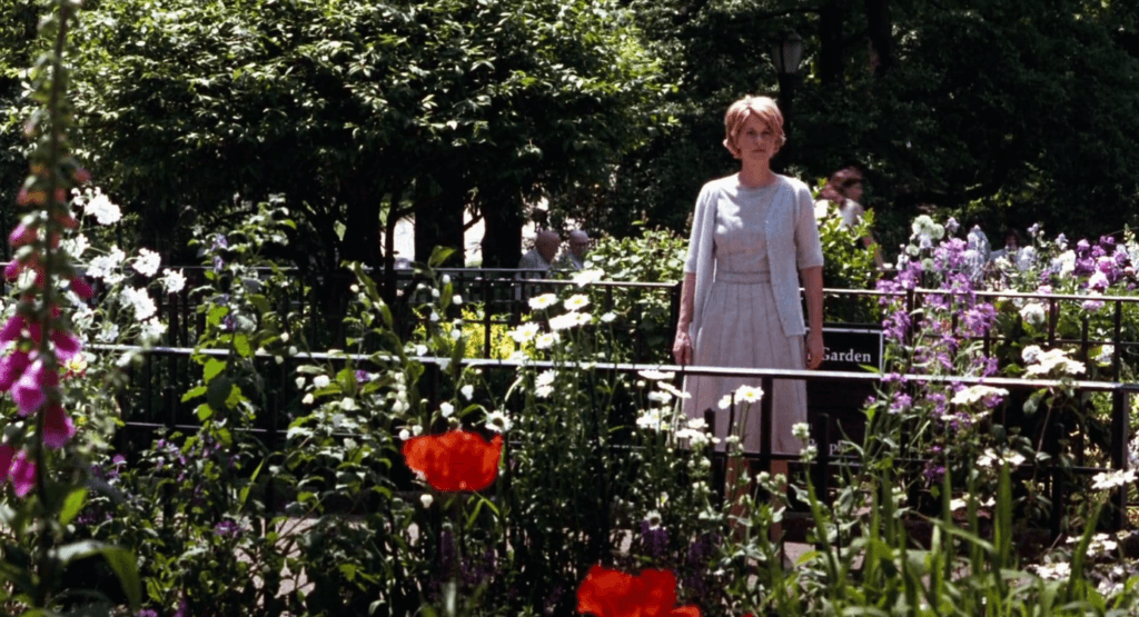 The lead female character wearing a spring blue dress and cardigan surrounded by flowers in a New York garden.