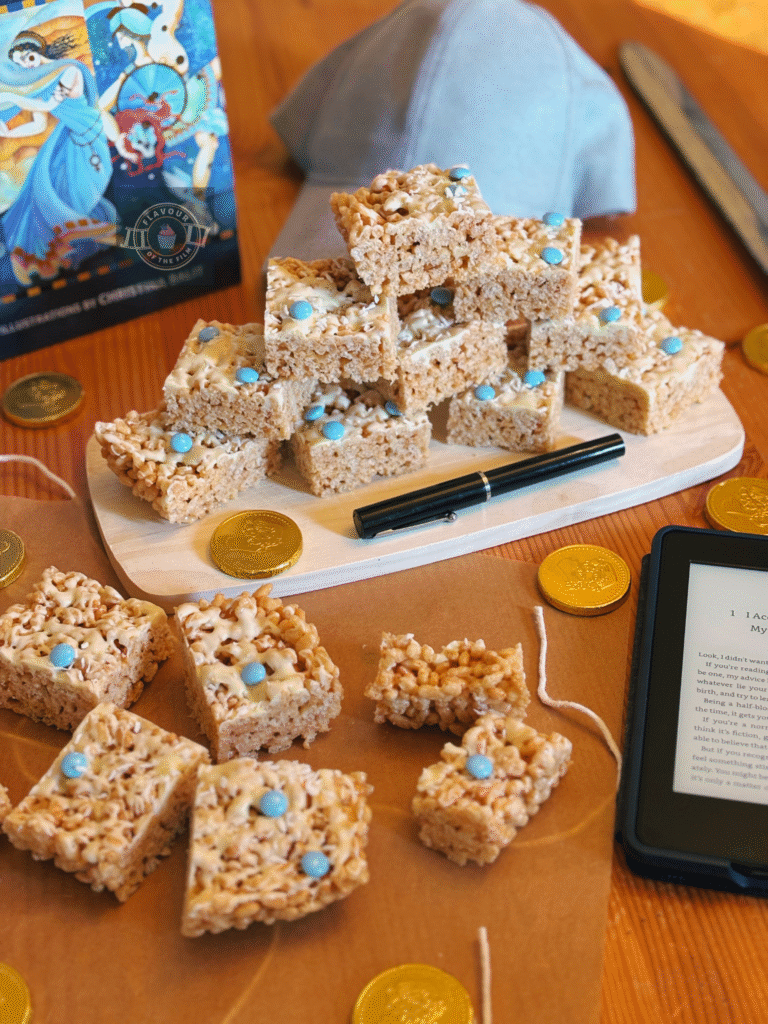 All images are of golden Percy Jackson ambrosia in the form of Rice Krispie treats which are topped with white chocolate, desiccated coconut and blue chocolate beans, and are displayed on a wooden board. Props include an illustrated copy of Greek mythology, a cap, a toy sword, a pen, a Kindle with the first page of Percy Jackson and the Lightning Thief, and gold coins.