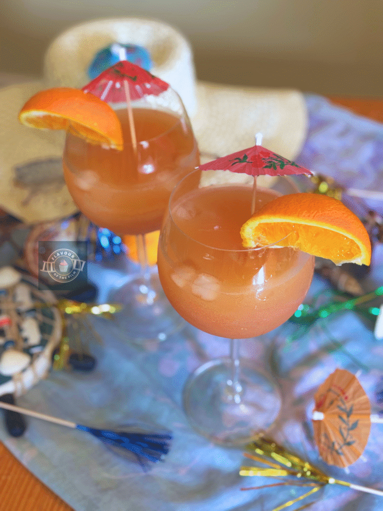 All images are of two glasses of the Mamma Mia! party mocktail, each with a wedge of orange on the rim of the glass and a pink drink umbrella. Props include a thin blue scarf, seashells, a sunhat, sunglasses and beach decor. Plus multi-coloured sparkler cocktail sticks.