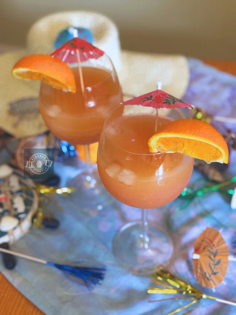 All images are of two glasses of the Mamma Mia! party mocktail, each with a wedge of orange on the rim of the glass and a pink drink umbrella. Props include a thin blue scarf, seashells, a sunhat, sunglasses and beach decor. Plus multi-coloured sparkler cocktail sticks.