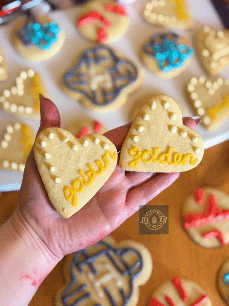 All images are of sugar cookies in round shapes, heart shapes, and four-rounded clover type shapes decorated with buttercream to represent aspects of K-Pop: Demon Hunters. The cookies are served on a purple and white board. Props include a sequin BTS cushion cover, a pink crochet turtle, a pink cat shaped fan, packets of ramyeon, tulip cups, and a pink and purple and blue disco ball pencil holder.
