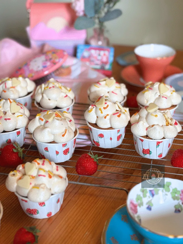 All images are of My Melody & Kuromi cupcakes in strawberry cupcake cases, topped with meringue clouds, white chocolate hearts, and sprinkles. Teh cupcakes are served on a wire rack. Props include pink oven gloves with peaches on, a Cinnamonroll fan, sunset bag, pink flowers in a pink vase, coral pink teacup and blue teacup, a Hello Kitty hairbrush, fresh strawberries, and a My Melody Amiibo card for Animal Crossing.